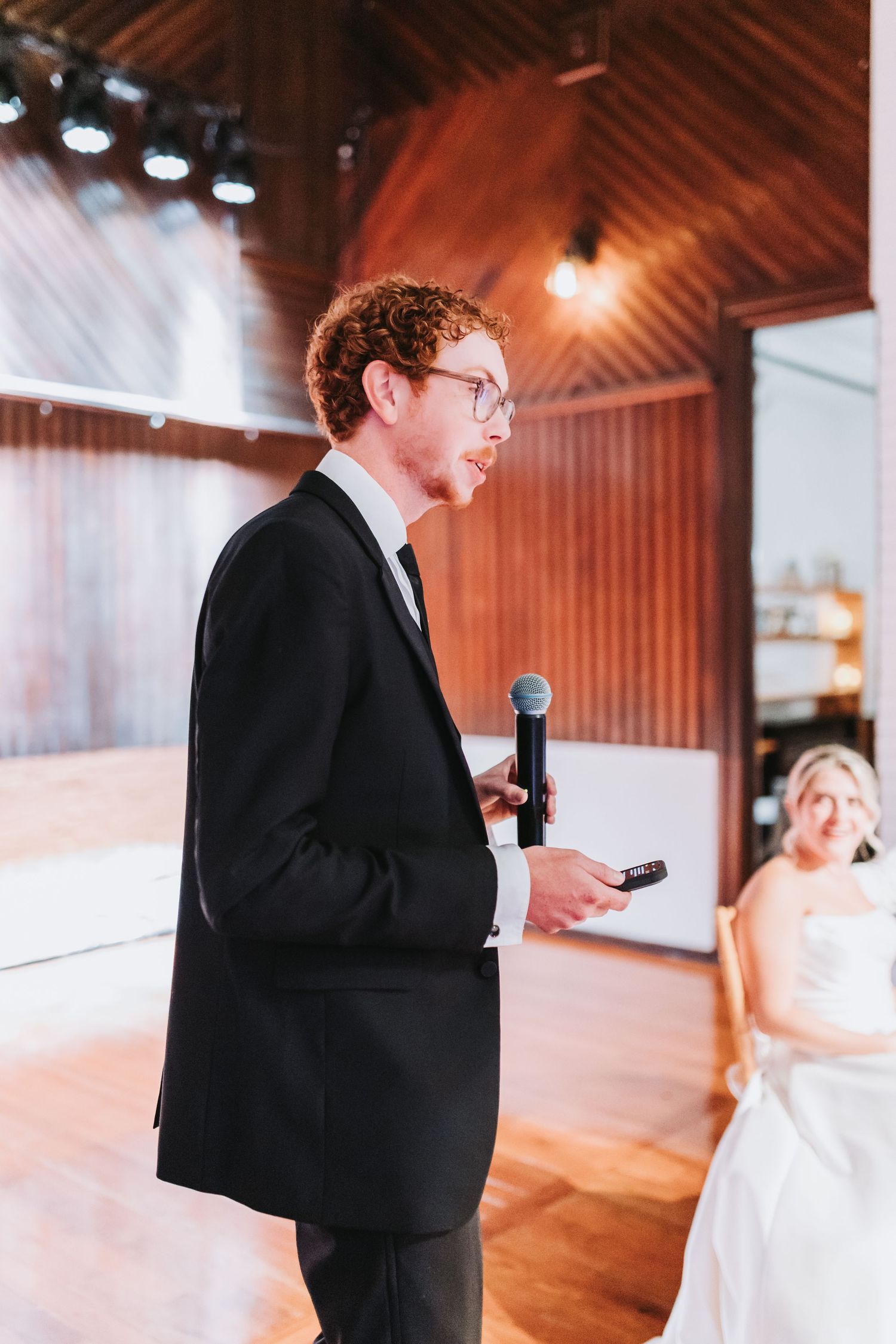 Curly red-haired man in tuxedo delivers a wedding toast speech with microphone in elegant venue.