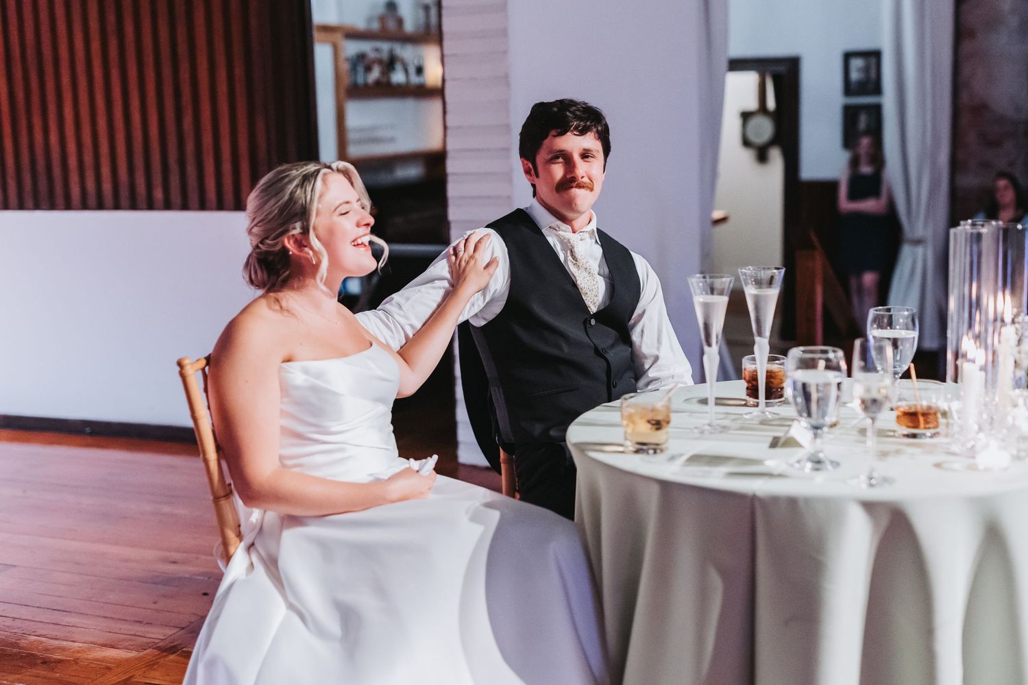 Bride and groom laugh together at sweetheart table during wedding reception.