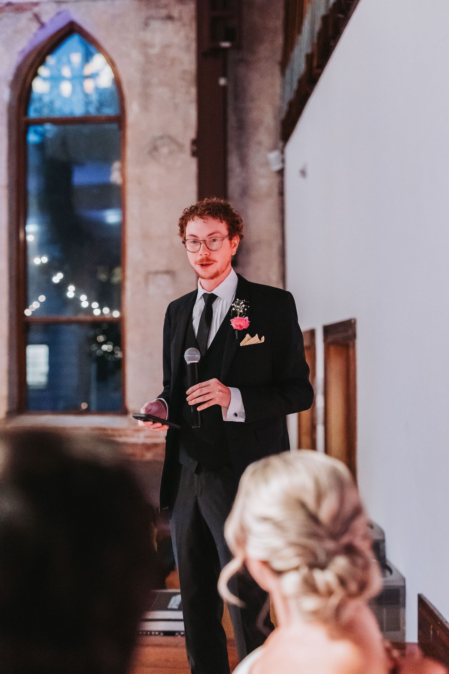 Dapper groomsman with curly hair delivers wedding speech holding microphone in church venue.