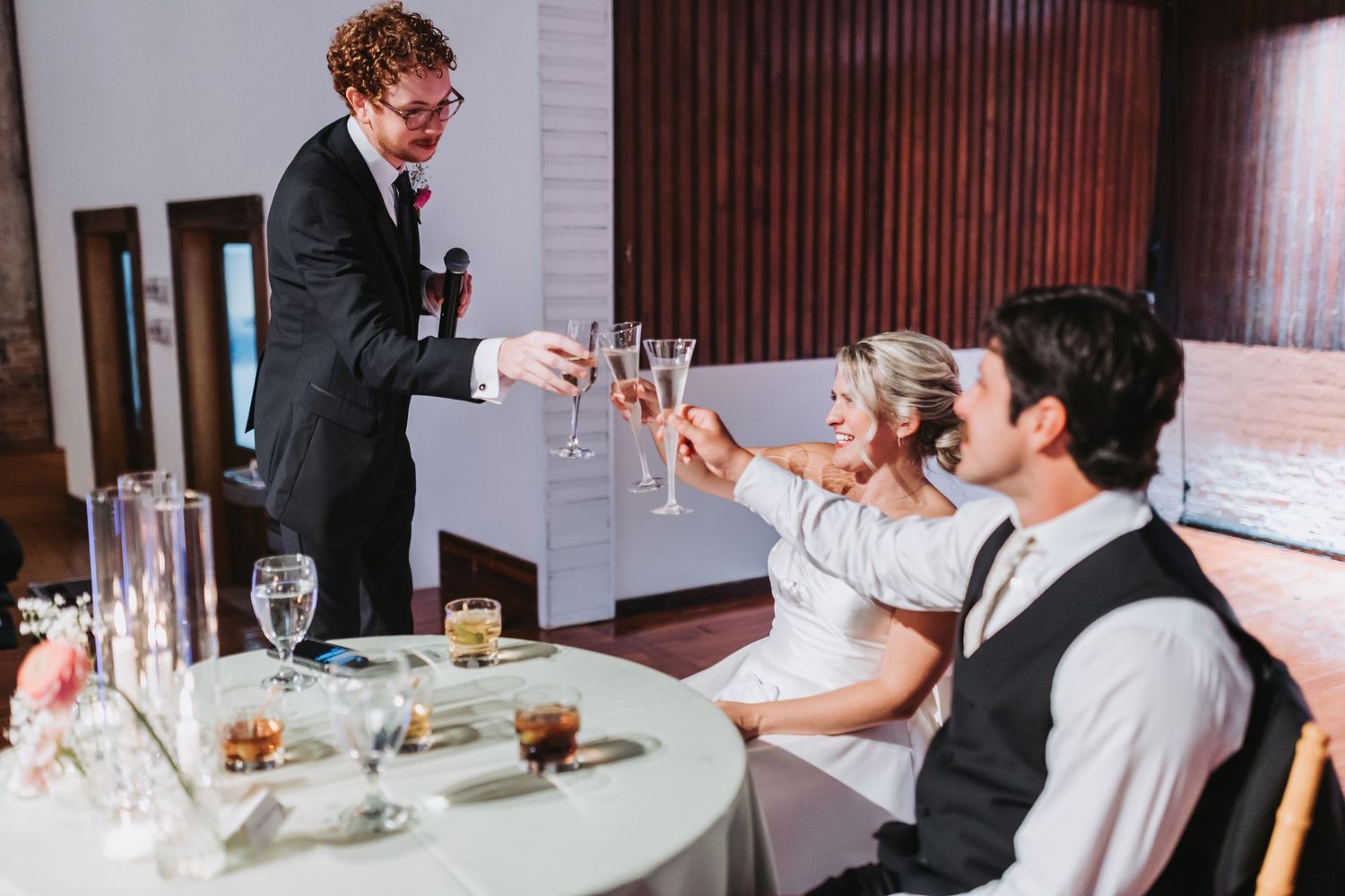 Best man toasts bride and groom with champagne glasses at wedding reception table.