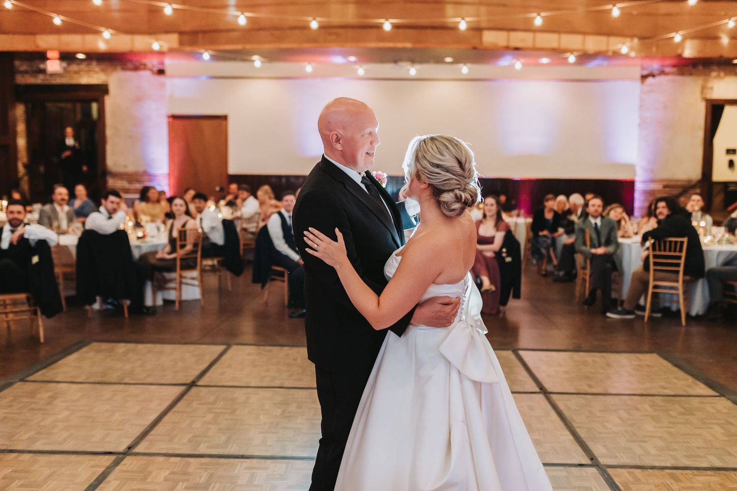 Bride and groom share romantic first dance on white dance floor with string lights at wedding reception.