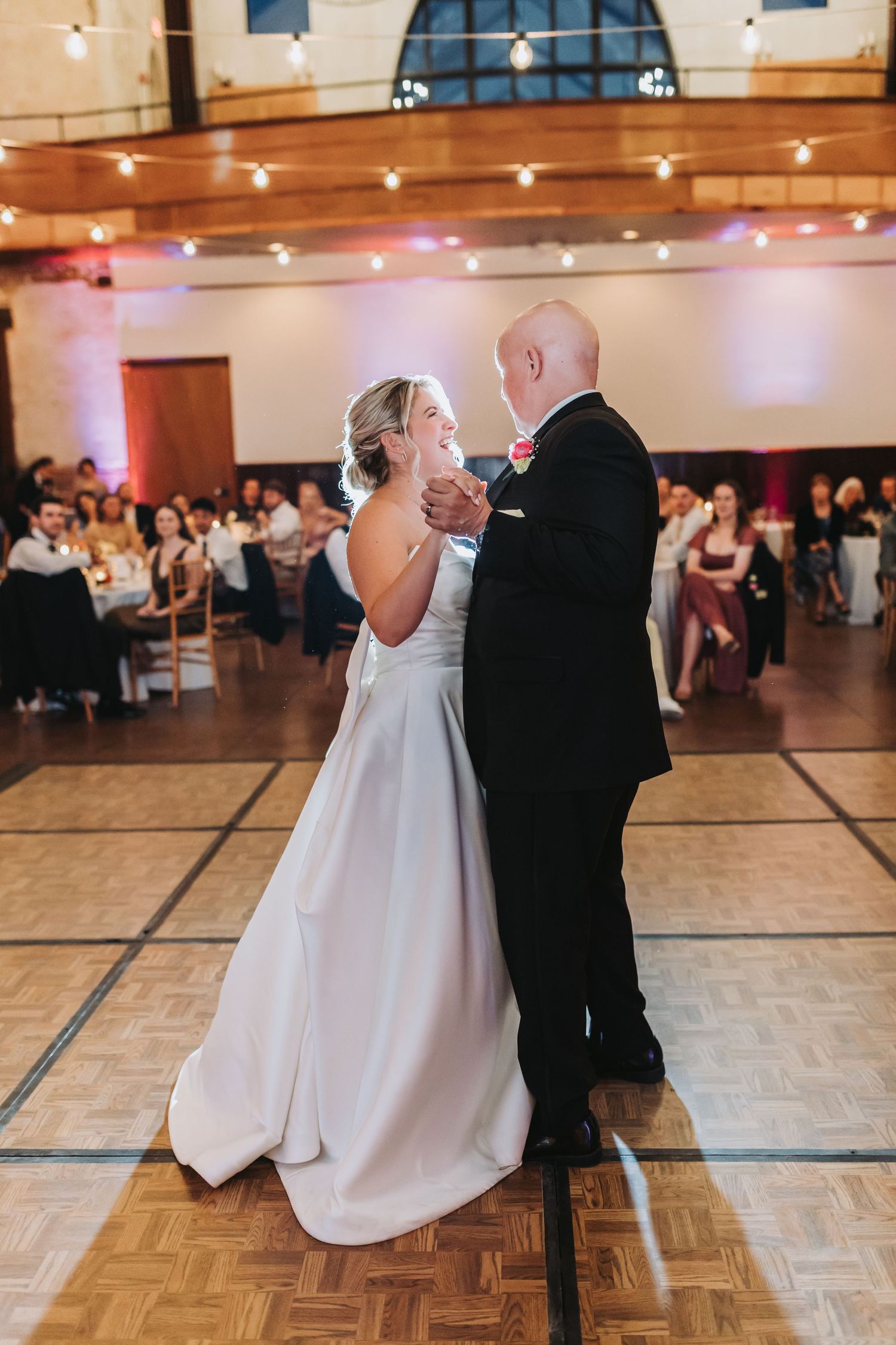 Bride in white gown shares a tender father-daughter dance on a wooden ballroom floor.