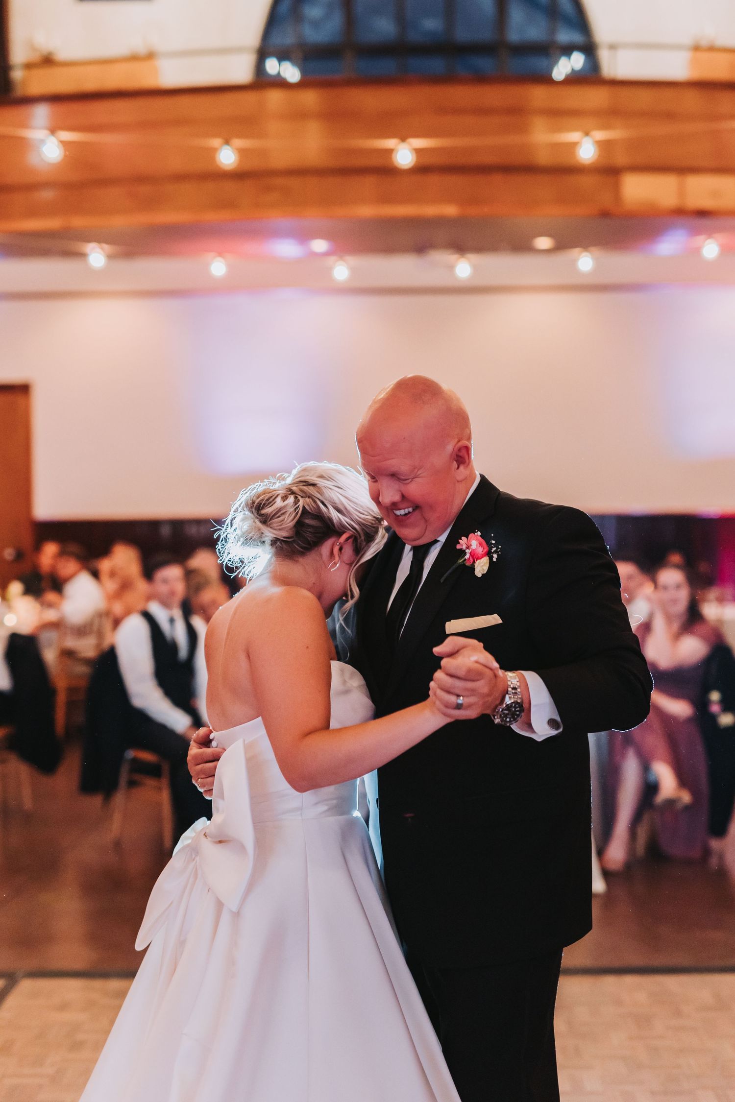 Bride and bald man in tuxedo share a laughing father-daughter dance at wedding reception.