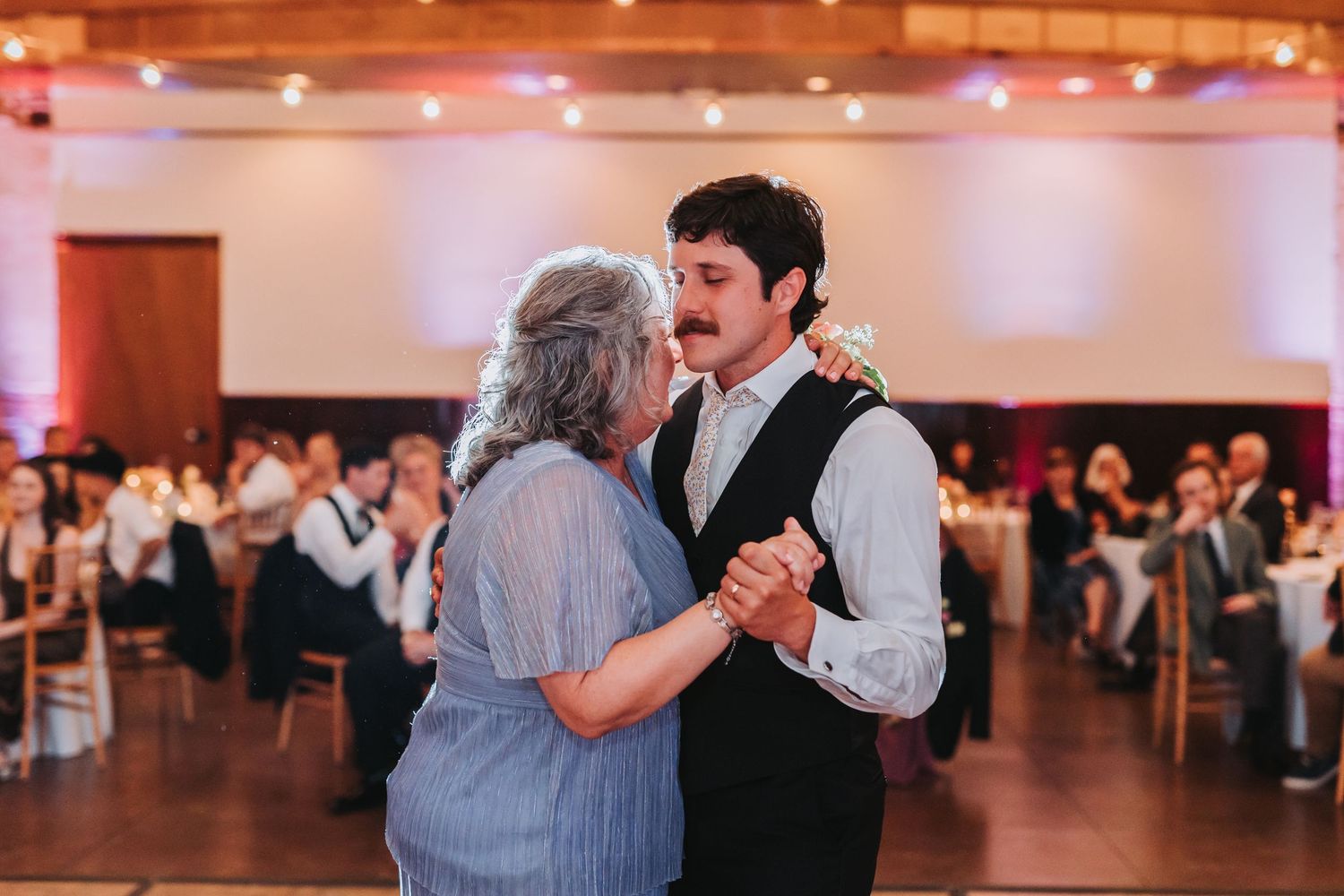 Groom dances with his mother during a heartfelt wedding reception moment in a decorated banquet hall.
