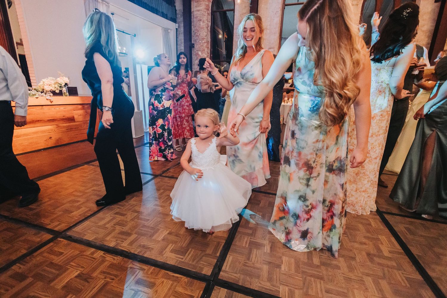 Adorable flower girl in white dress dances with women in floral gowns at wedding reception.