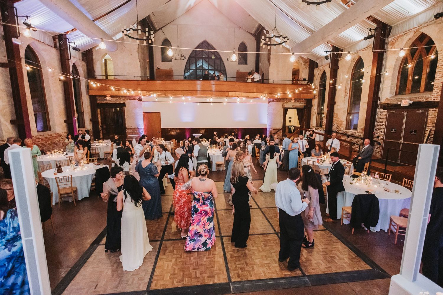 Guests fill a grand church wedding reception hall with string lights and wooden dance floor.