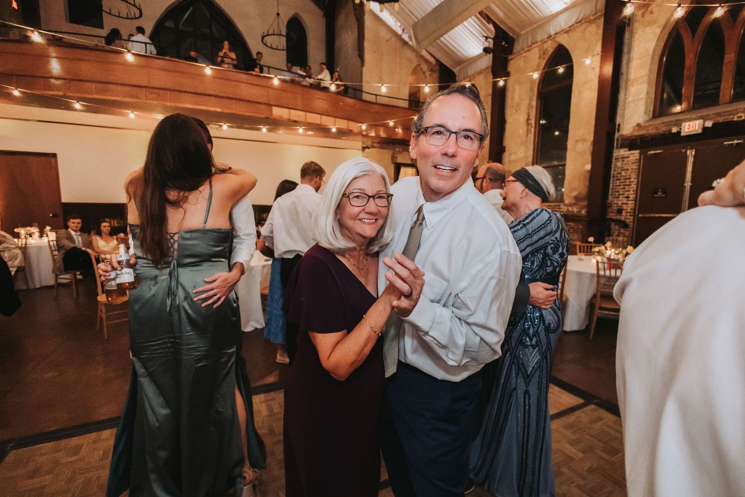 Couple smiling and dancing together at an elegant wedding reception in a historic venue with string lights.