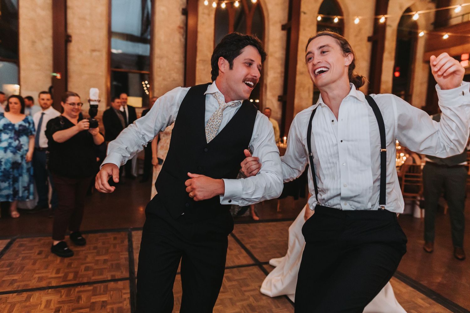Two guests in suspenders laugh and dance energetically on the wedding reception dance floor.