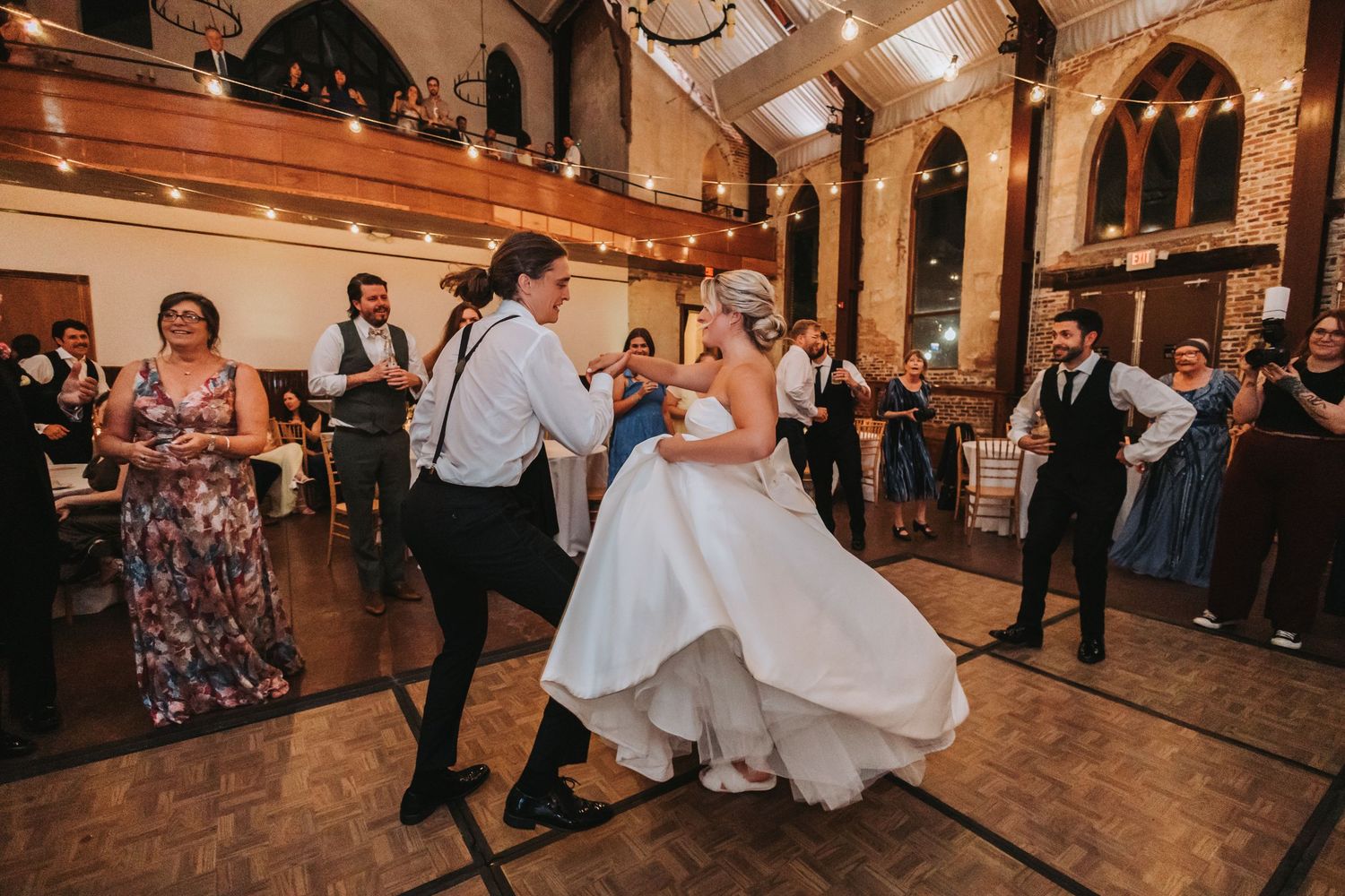 Bride and groom share energetic first dance at wedding reception in a stunning church venue with string lights.