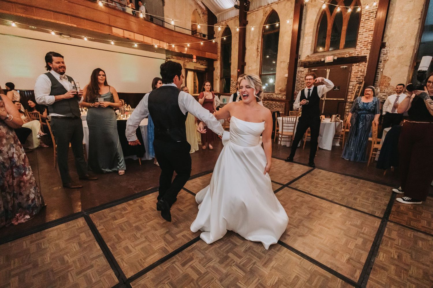 Bride and groom share their first dance on a wooden dance floor surrounded by guests at their wedding reception.