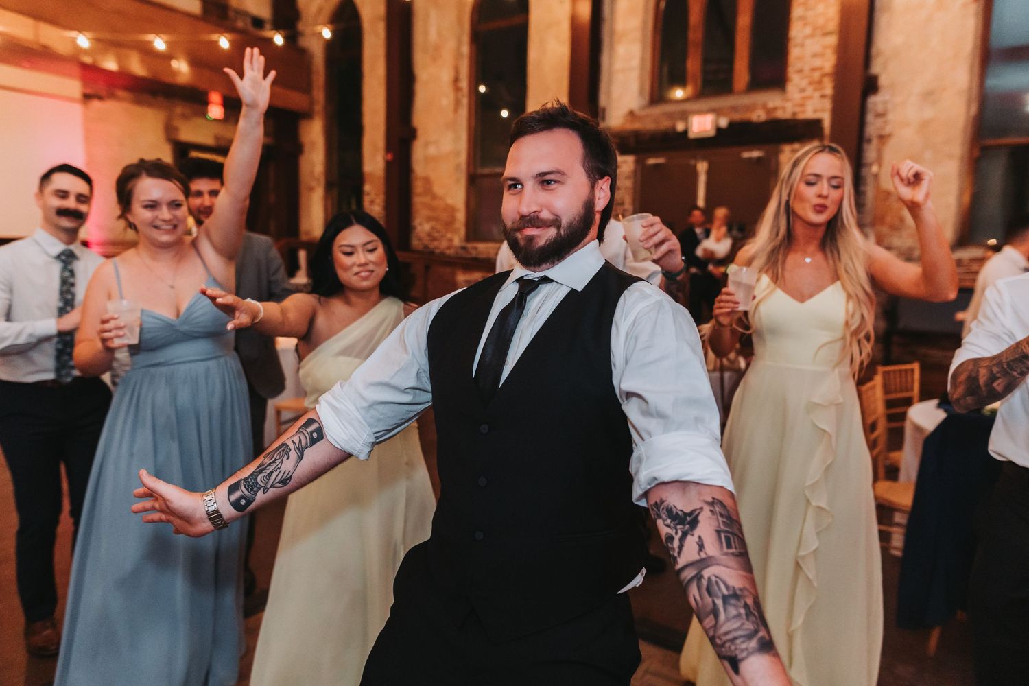 Tattooed groomsman dances energetically with bridesmaids and guests during wedding reception party.