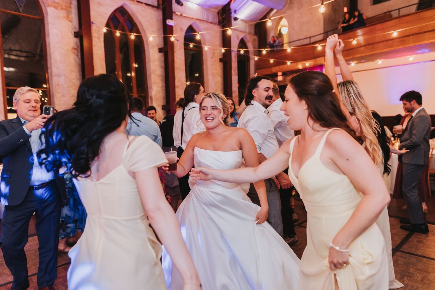Bride dancing joyfully with bridesmaids in white dresses at a wedding reception with string lights.