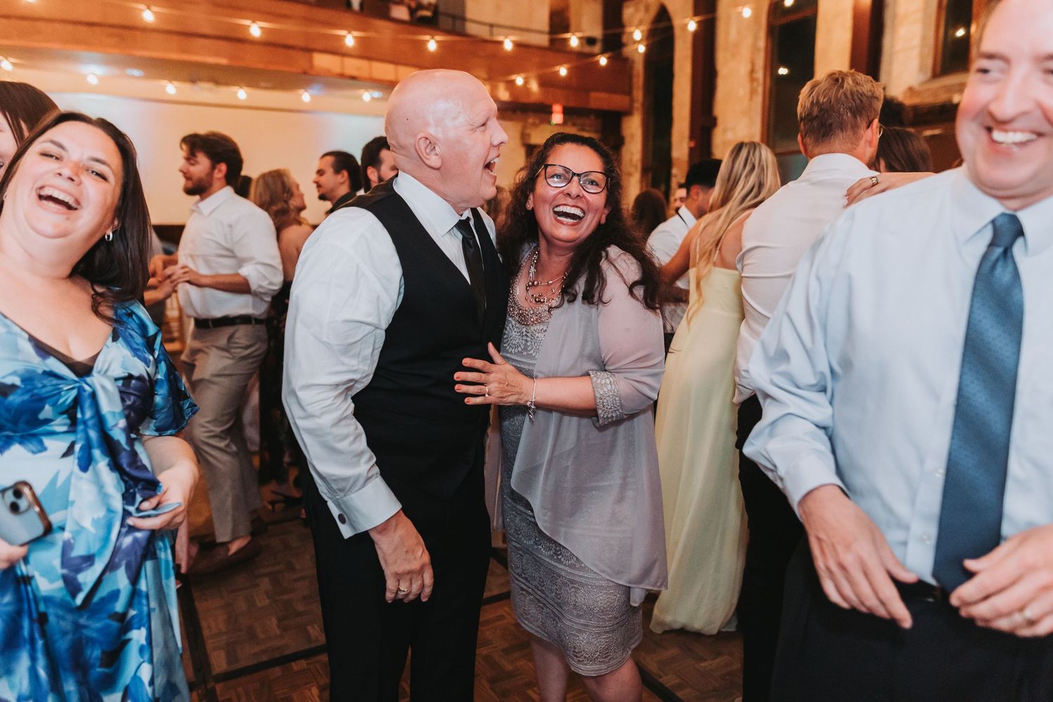 Joyful wedding guests laughing and dancing together at a festive indoor reception with warm string lights.