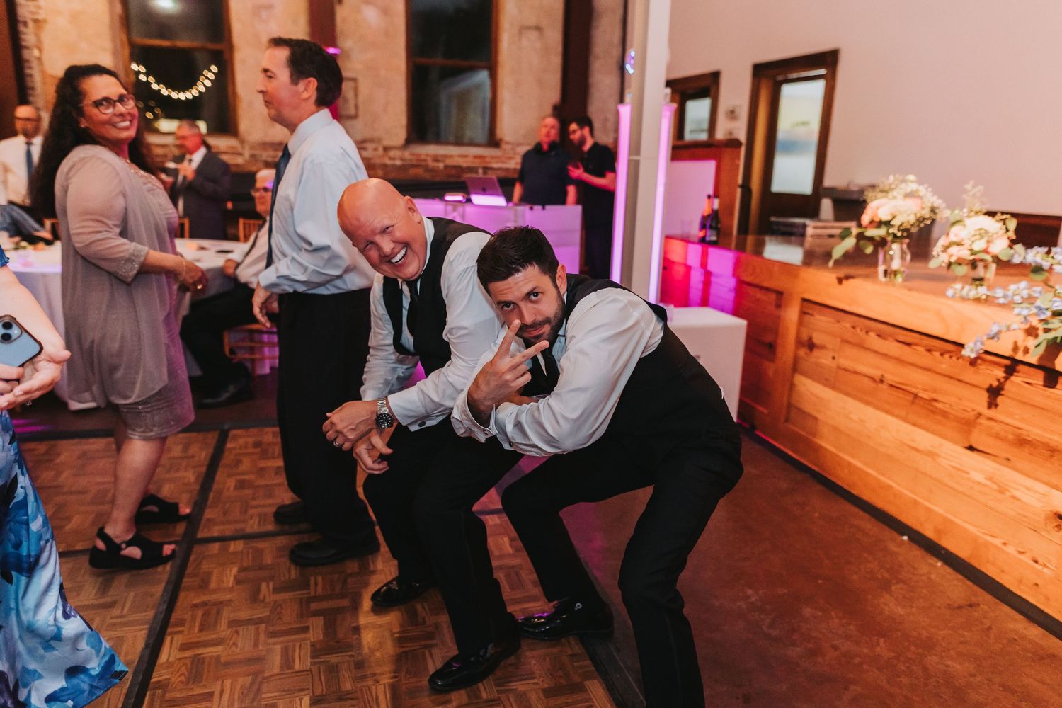 Two men strike fun poses on a wedding reception dance floor while guests mingle in the background.