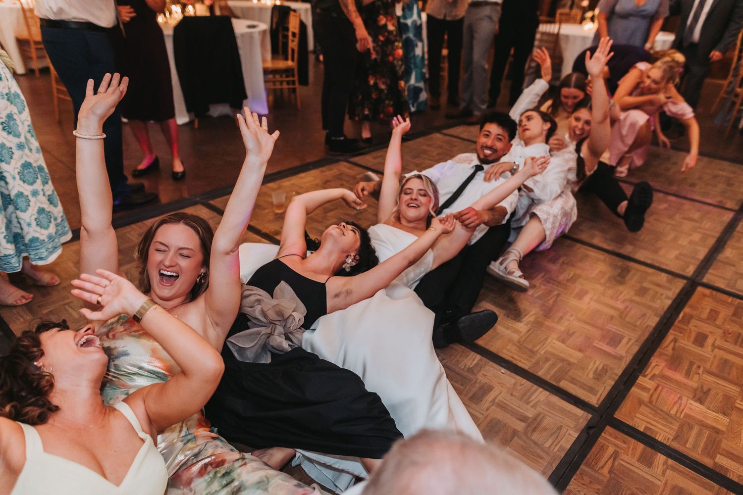 Wedding guests laughing and sitting on dance floor during lively reception celebration.