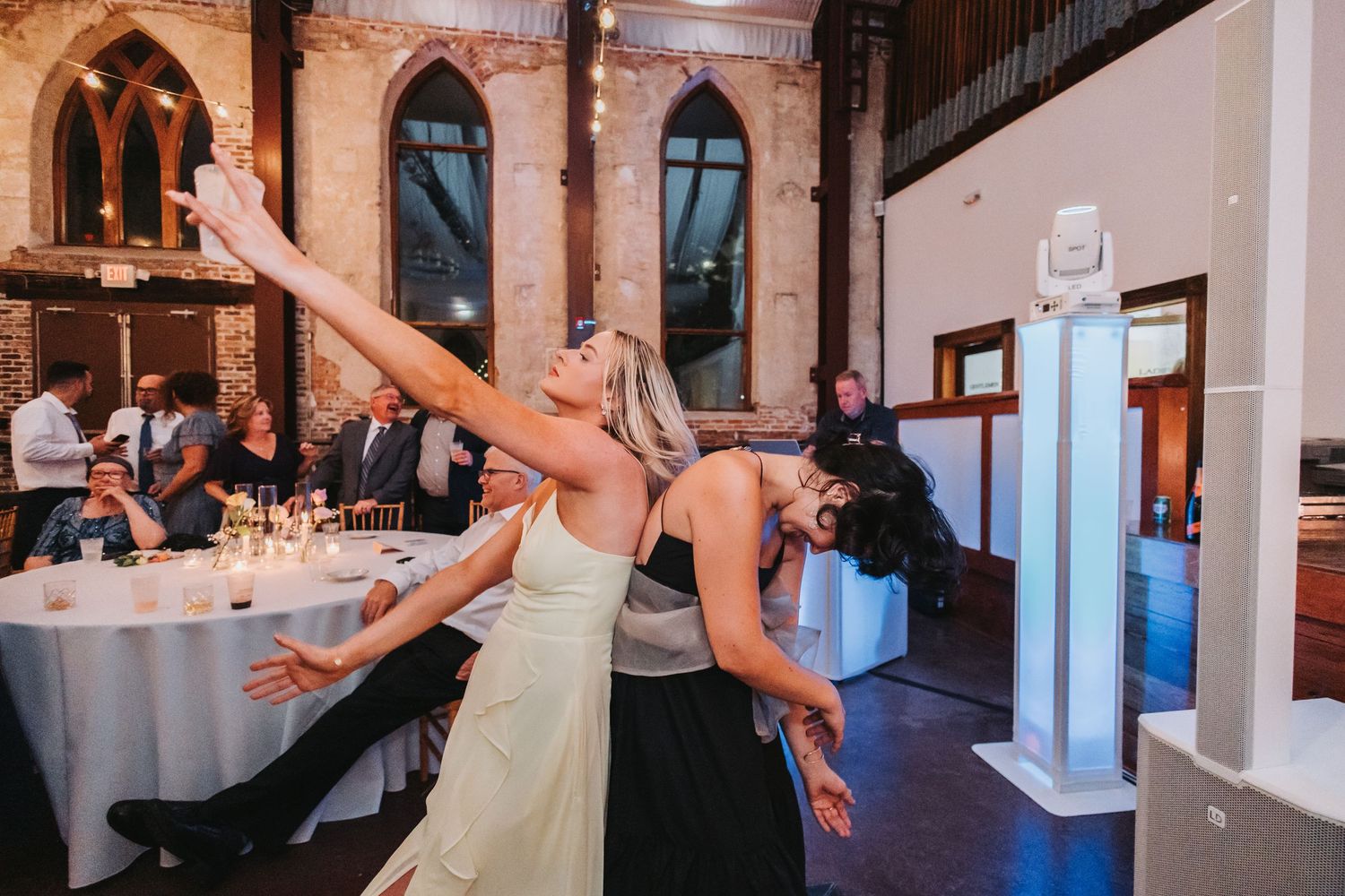 Two women dancing joyfully at a wedding reception in an elegant venue with arched windows and guests in background.