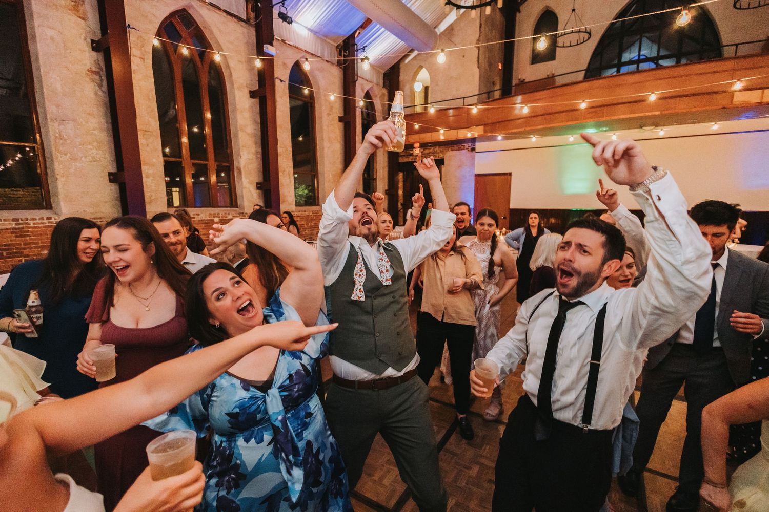 Guests dancing joyfully at a wedding reception in a decorated venue with string lights overhead.
