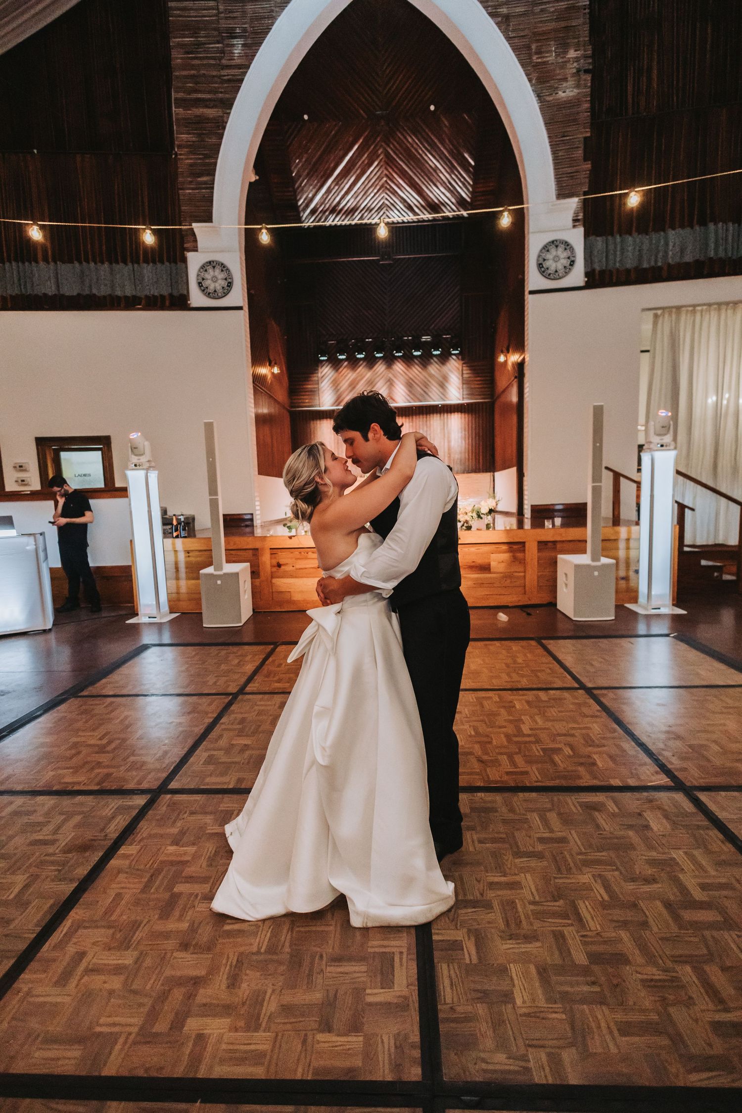 Bride and groom share their first dance on a wooden dance floor inside an elegant wedding venue with arched ceilings.