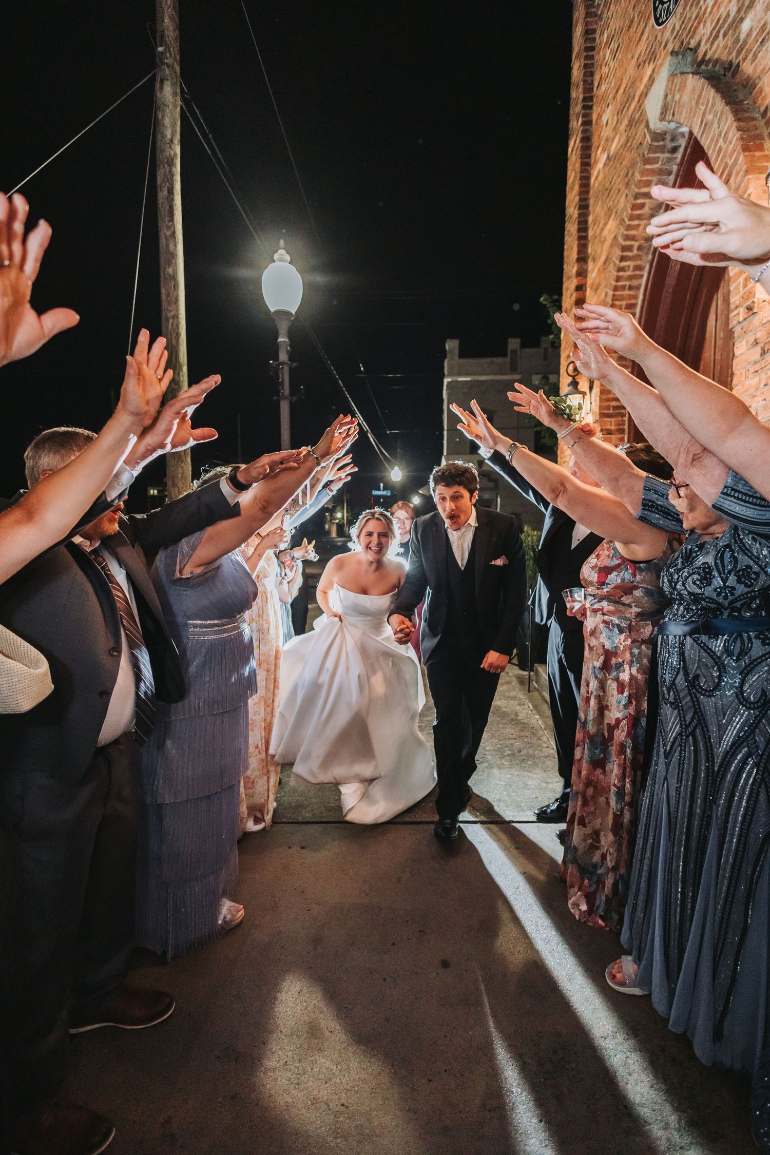 Bride and groom exit through a tunnel of cheering guests waving their hands at a nighttime wedding sendoff.