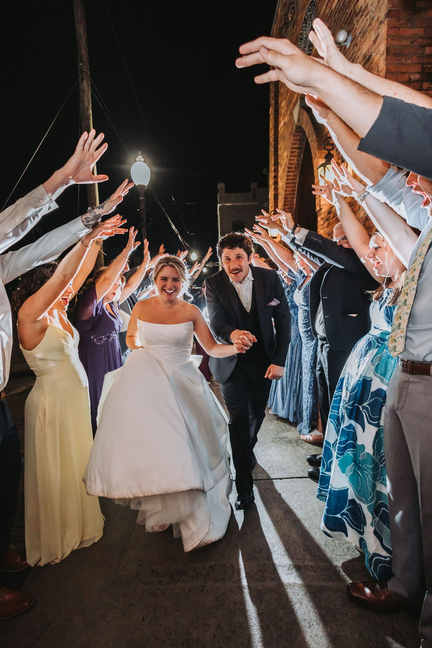 Bride and groom walk through sparkler tunnel with cheering guests lining the path during outdoor wedding night sendoff.