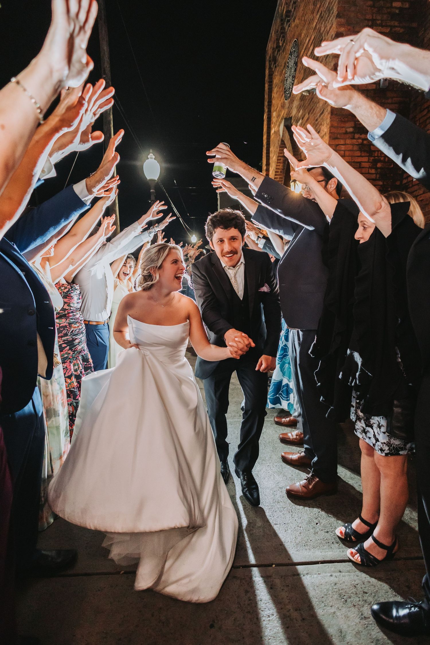 Bride and groom exit wedding reception through sparkler tunnel surrounded by cheering guests at night.