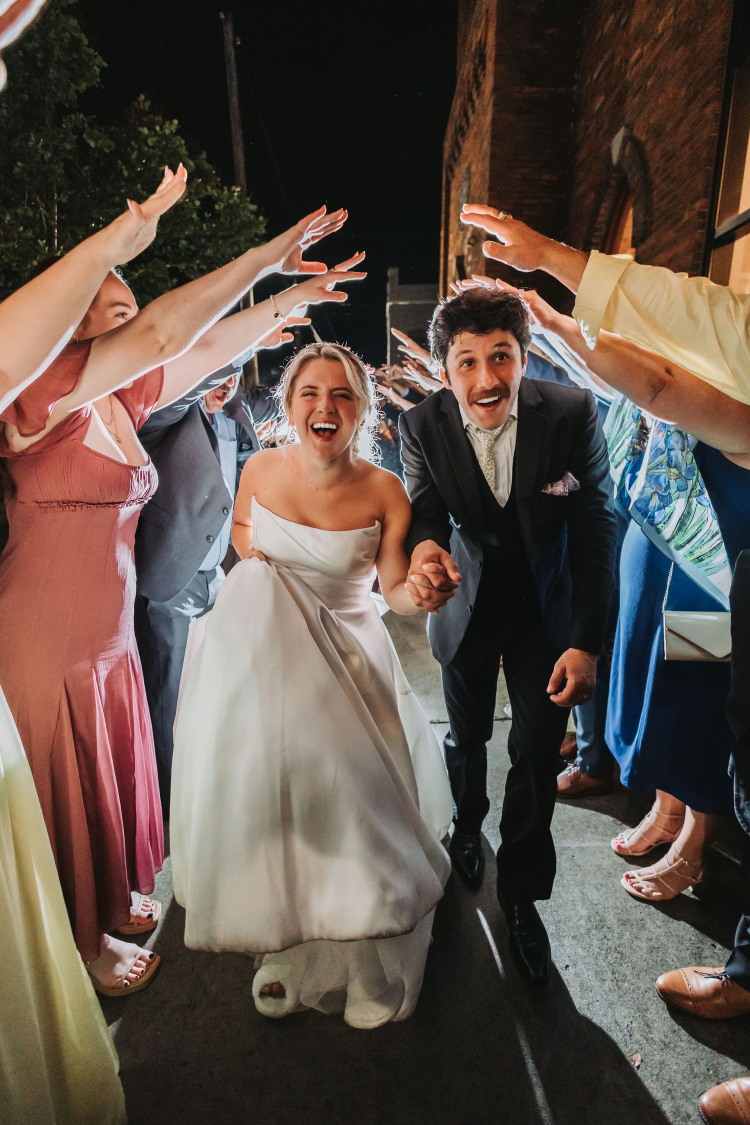 Joyful newlyweds run through a tunnel of raised hands from wedding guests during their celebratory send-off at night.