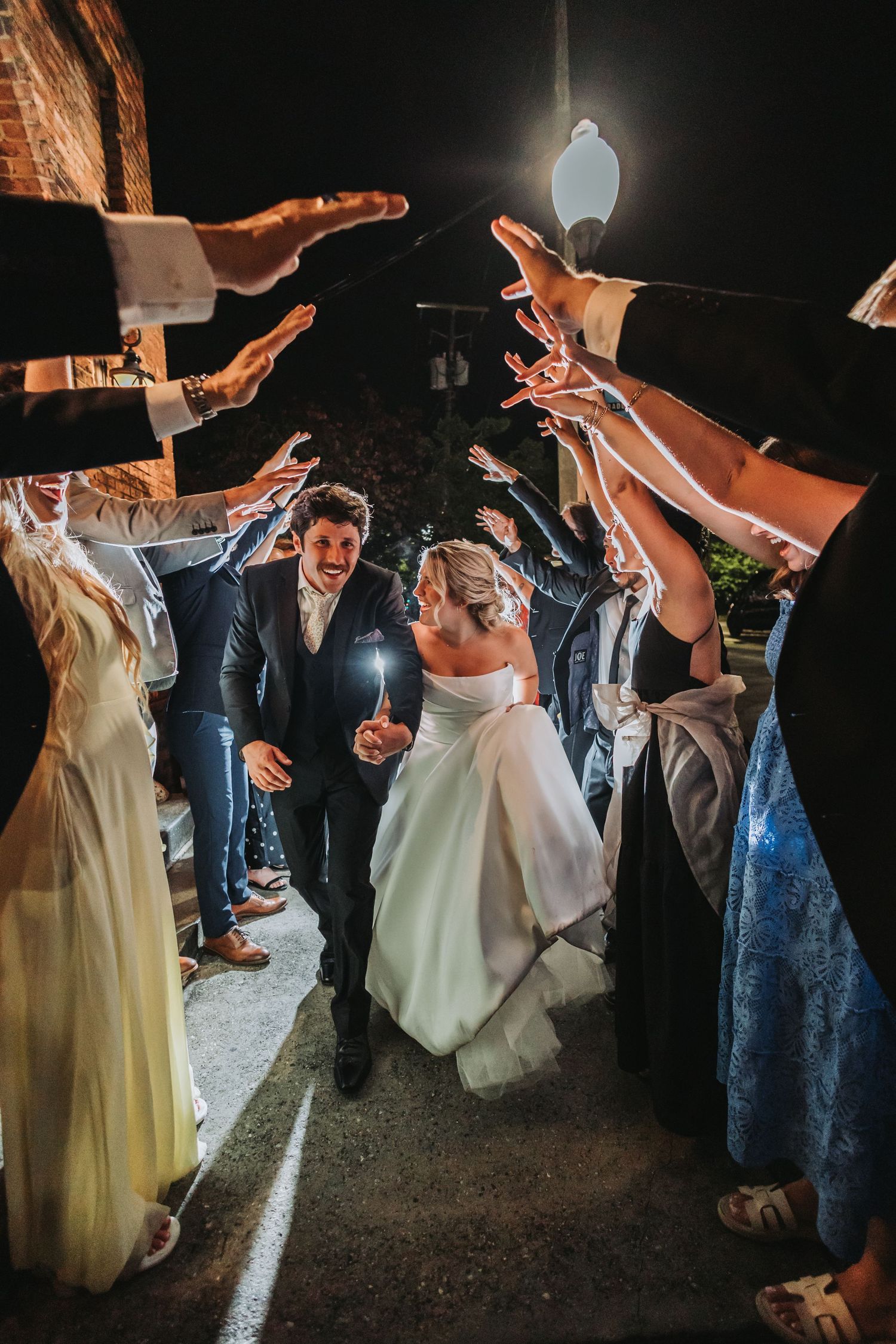 Bride and groom walk through a sparkler tunnel of cheering wedding guests at night.