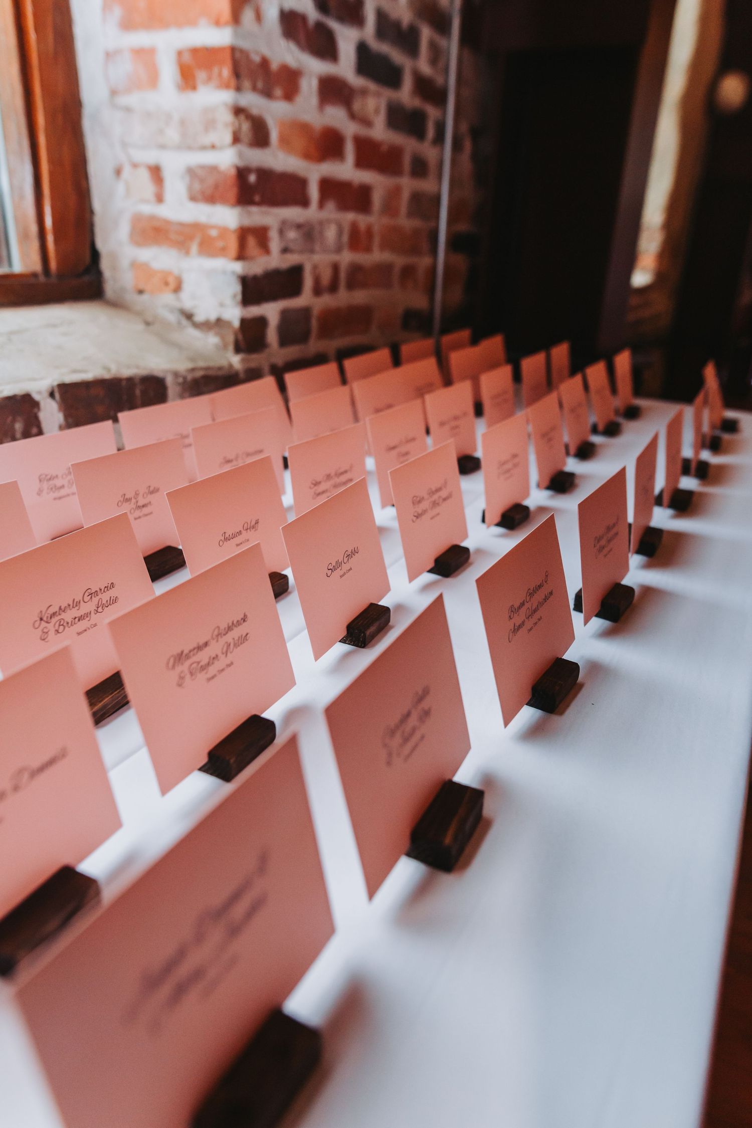 Rows of pink escort cards displayed in wooden holders on a white table against a rustic brick wall.