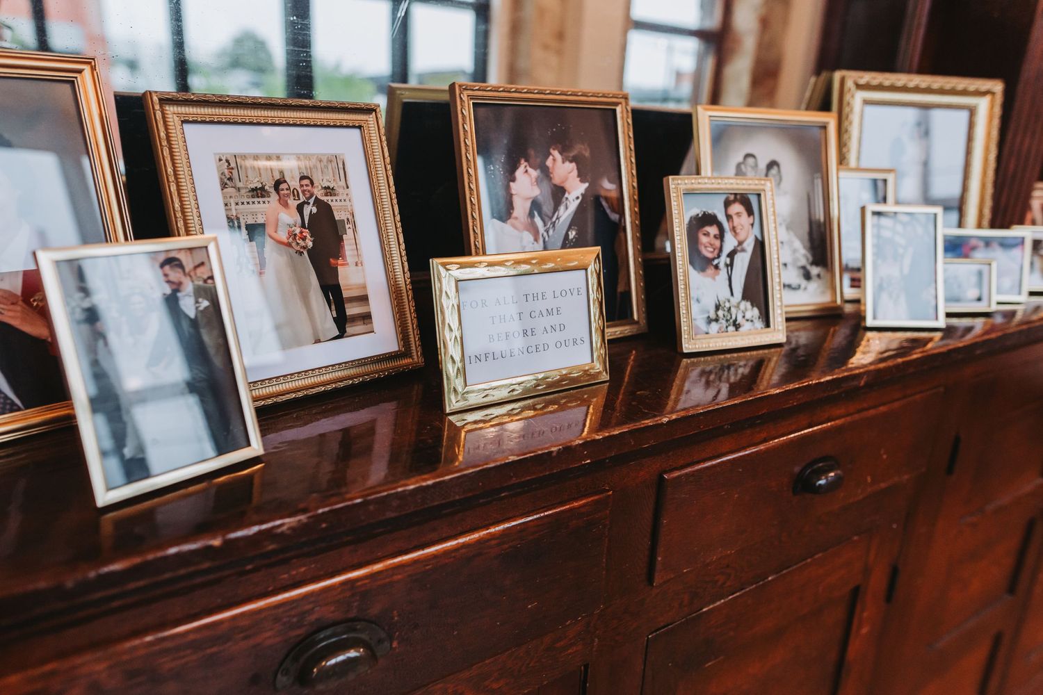 Gold-framed wedding photos displayed on a dark wood dresser at a vintage-inspired wedding reception venue.