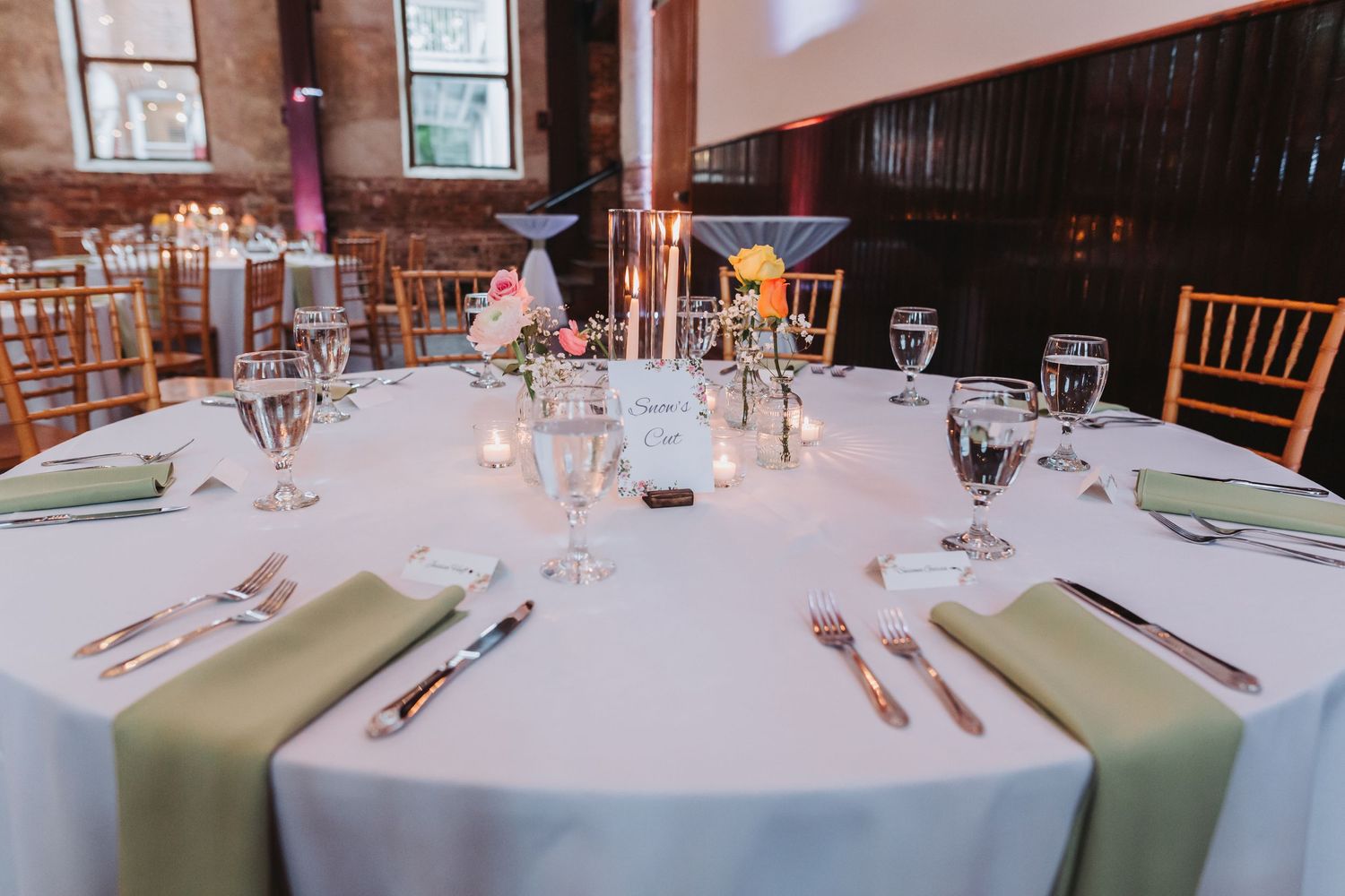 Elegant wedding reception table with sage green napkins, crystal glassware, candles, and floral centerpiece in a rustic venue.
