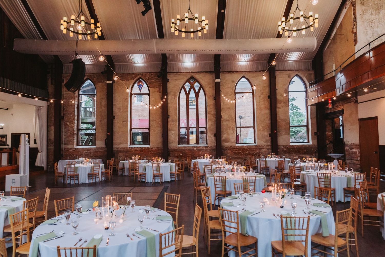 Elegant wedding reception hall with gothic windows, round tables, and warm chandelier lighting.