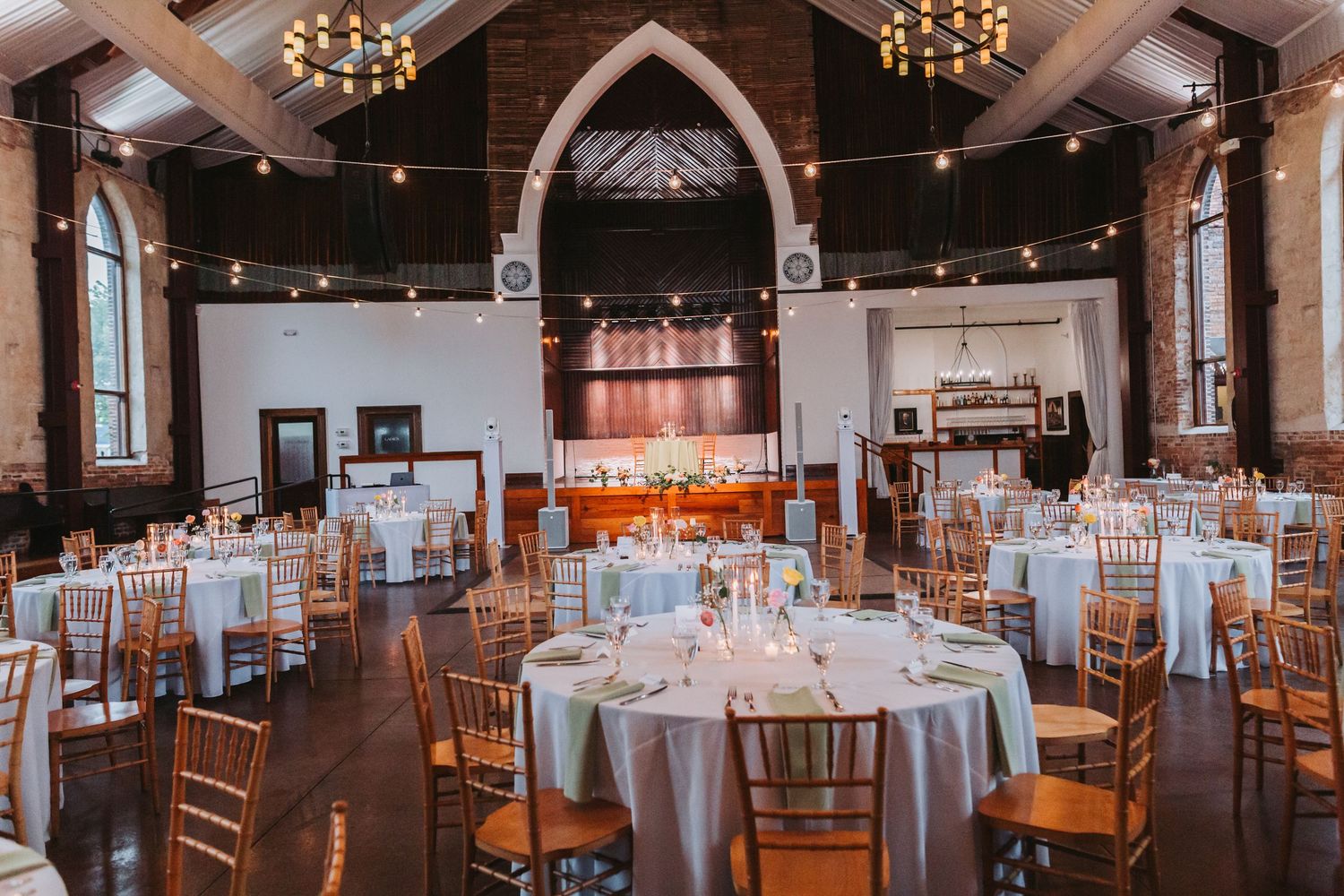 Elegant wedding reception in a historic venue with string lights, round tables, chiavari chairs, and arched windows.