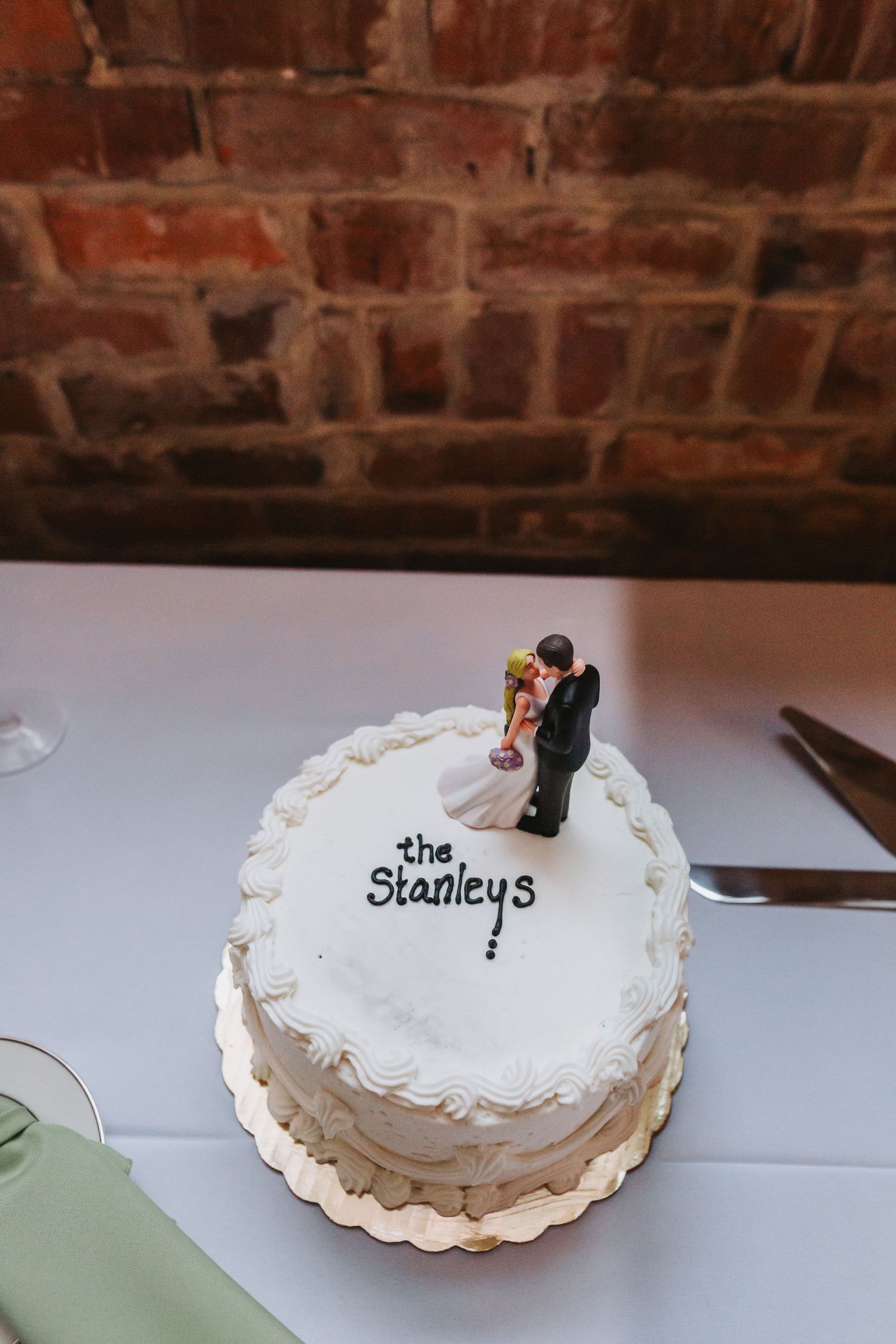 White wedding cake with bride and groom topper reading The Stanleys against a brick wall backdrop.
