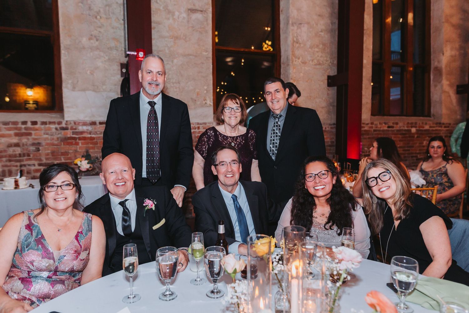 Group of seven people posing at a formal event dinner table with candles and glassware in elegant venue.