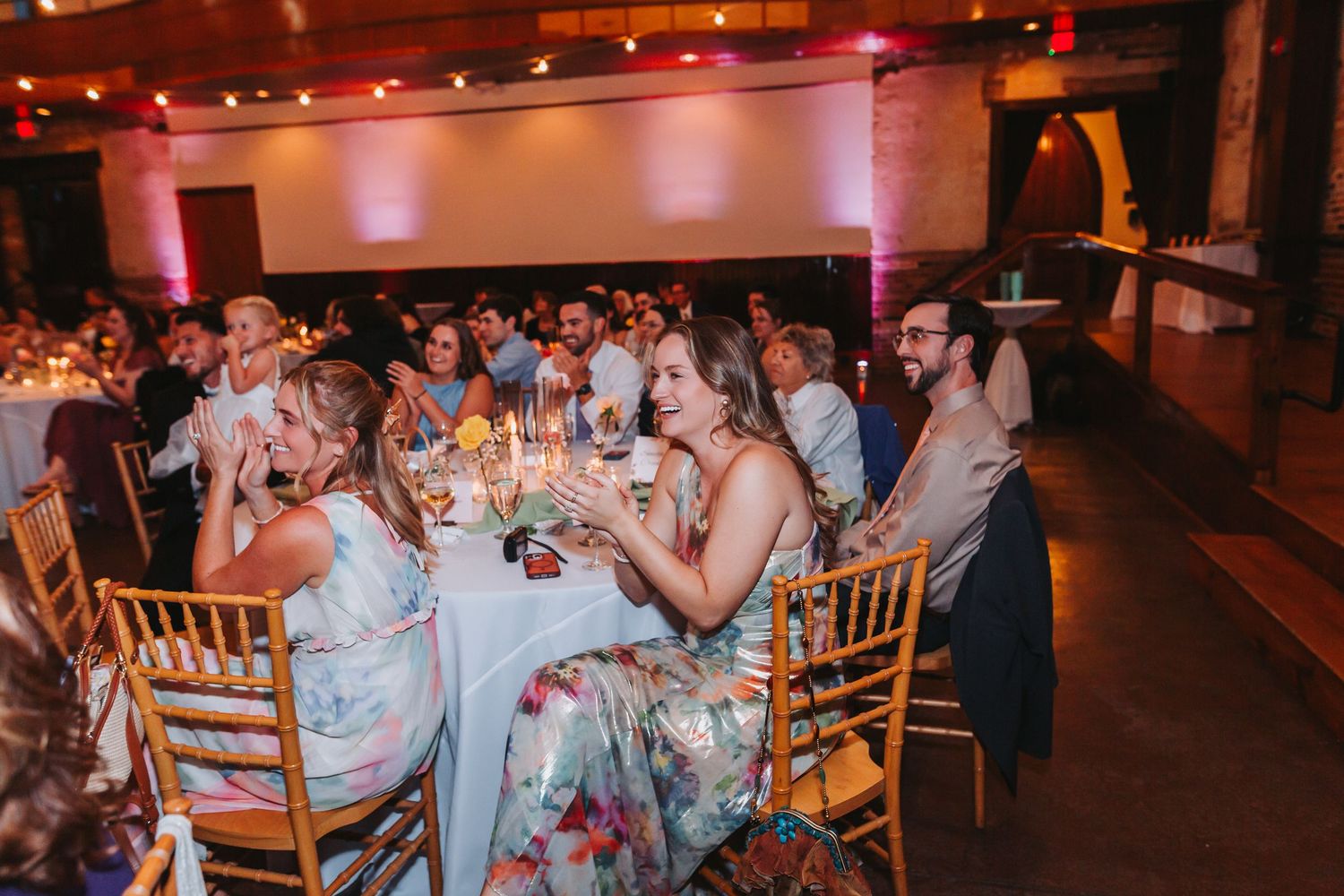 Guests laughing and celebrating at a wedding reception dinner in an elegant venue with warm pink lighting.