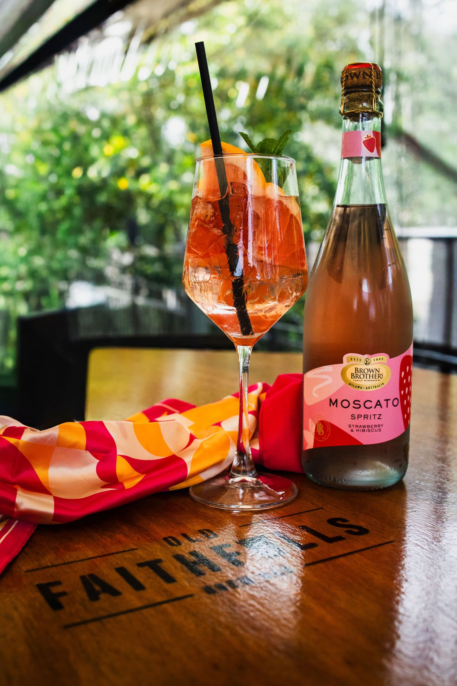 Brown Brothers Moscato Spritz bottle beside a chilled orange spritz cocktail with a black straw on a wooden table with a colorful napkin.