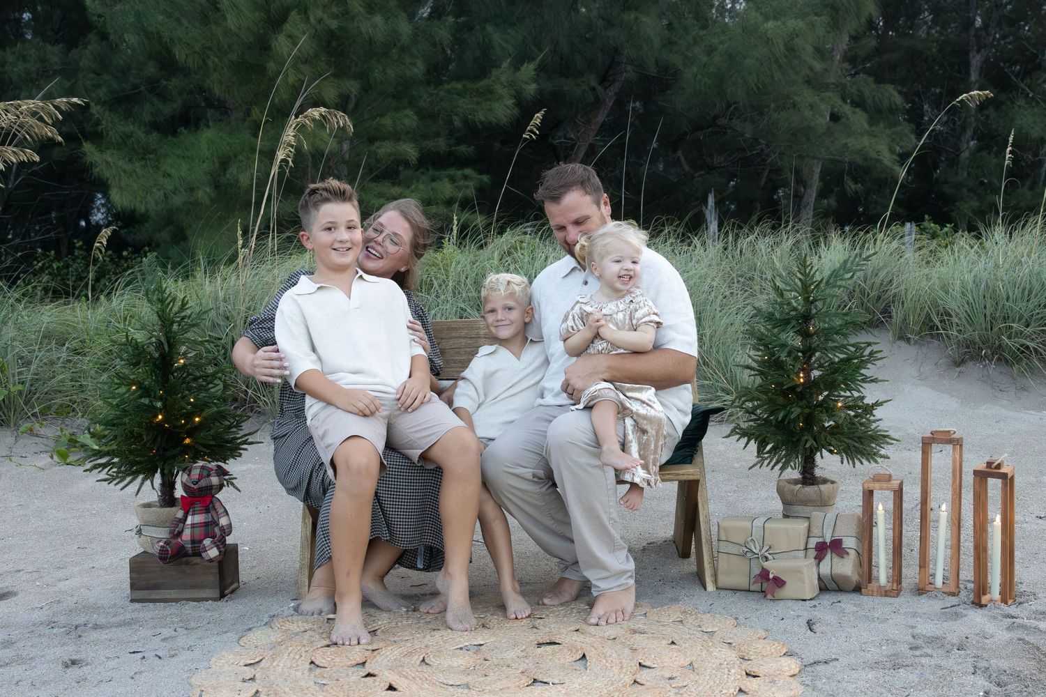 Smiling family of five sits together on a sandy beach beside small decorated Christmas trees and wooden lanterns for a holiday portrait.