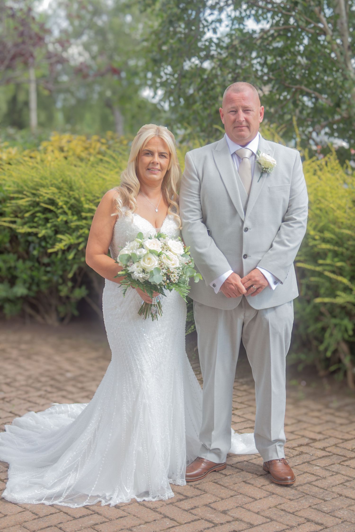 Smiling bride in a white lace wedding gown holding a bouquet stands beside a groom in a light gray suit on a brick path by green hedges.