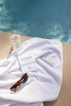 packaging oslo skin lab posé sur une serviette de plage avec des lunettes de soleil et un verre d'eau au bord d'une piscine