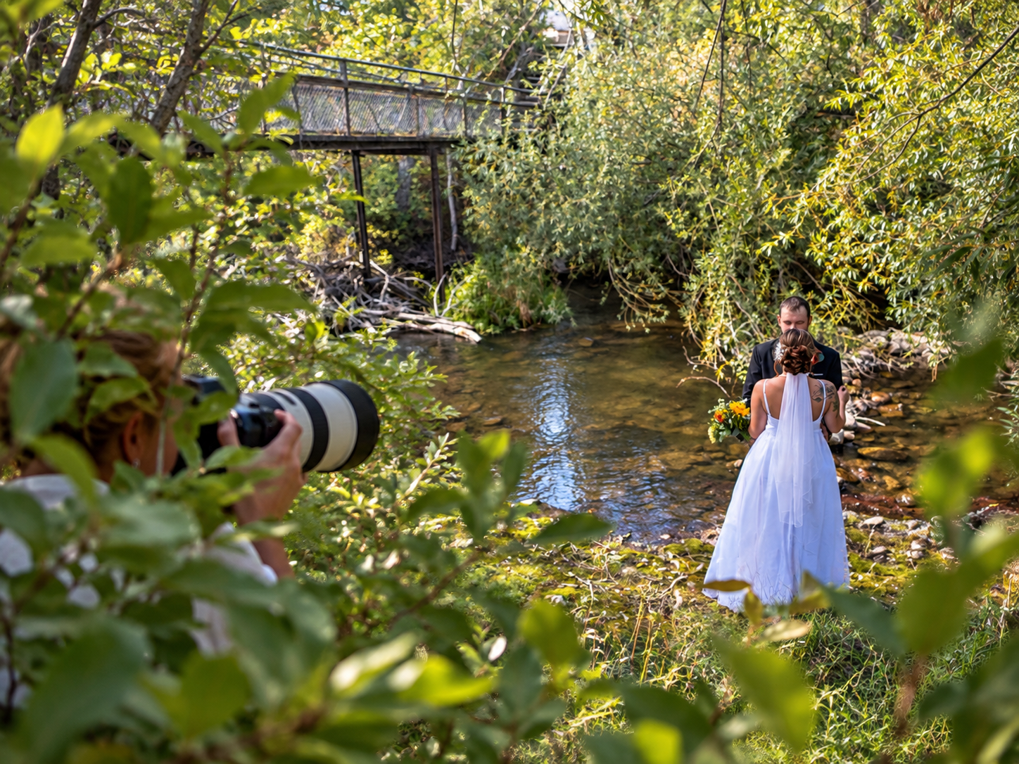 Photographer capturing bride in white gown by a serene creek surrounded by lush green trees and foliage.