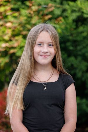 Young girl with long blonde hair wearing a black top and star necklace smiling outdoors.