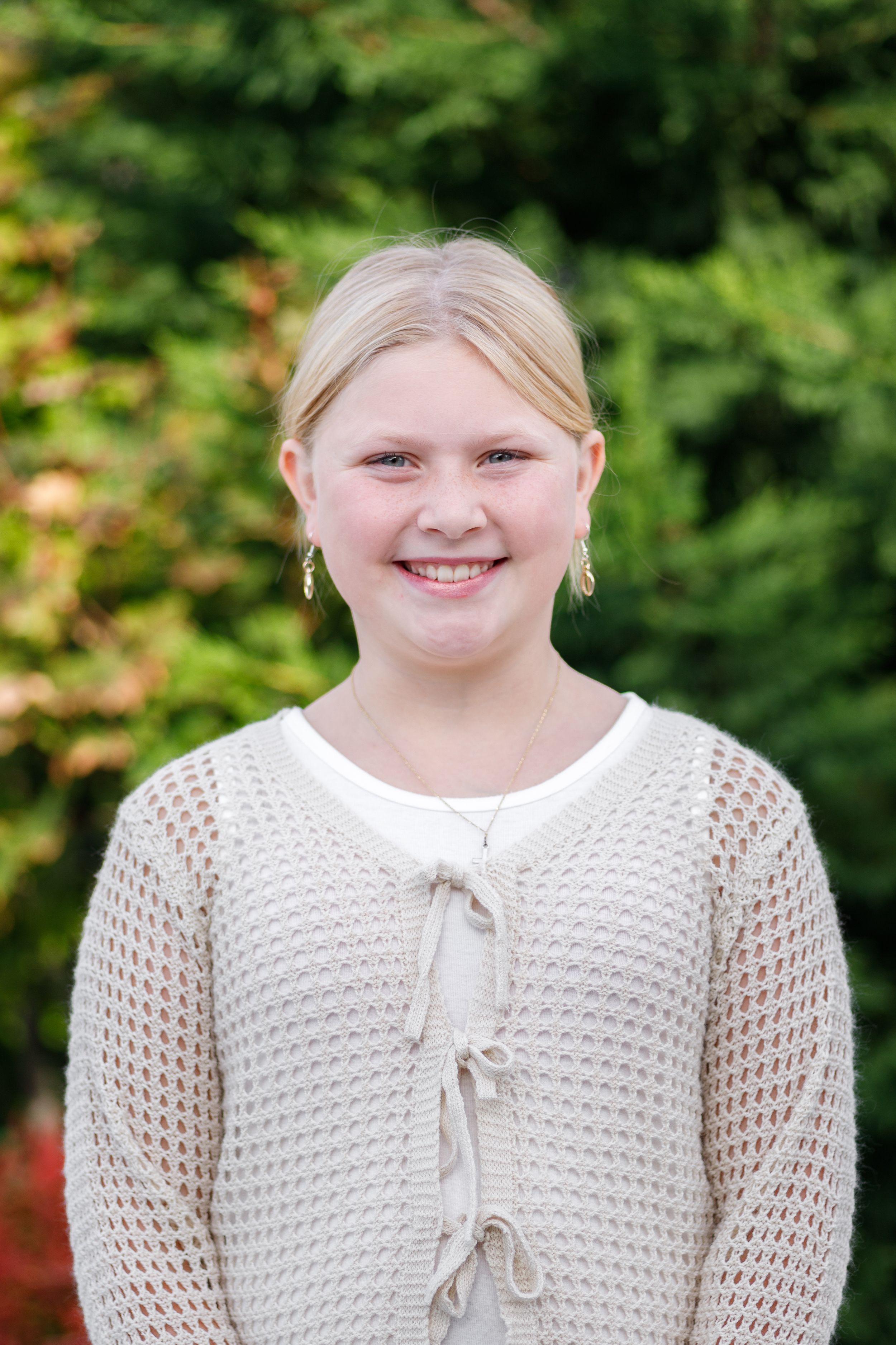 Smiling blonde girl wearing a cream knit cardigan and necklace, posing outdoors with green trees in background.