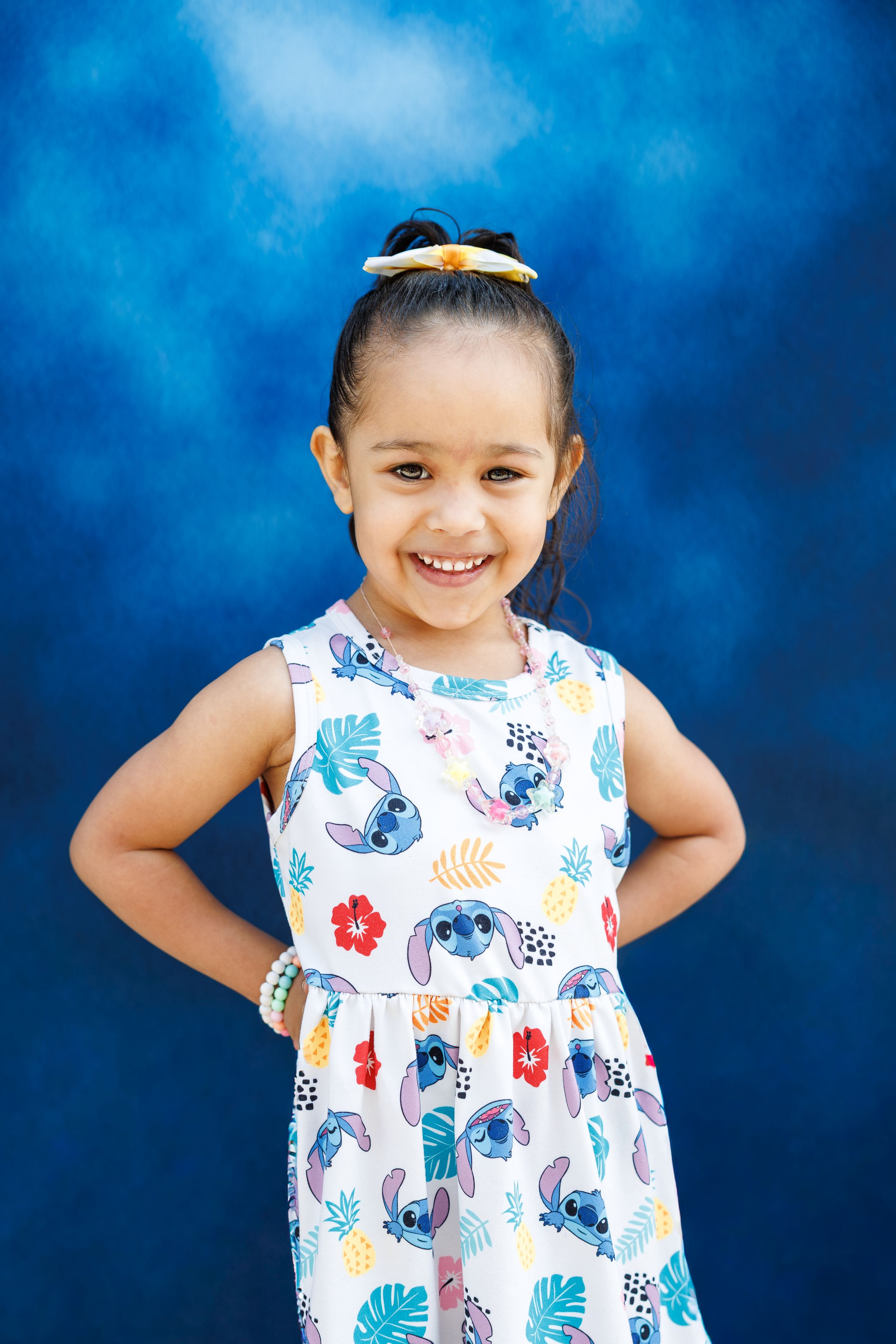 Smiling young girl in a Stitch print dress posing with hands on hips against a blue background.