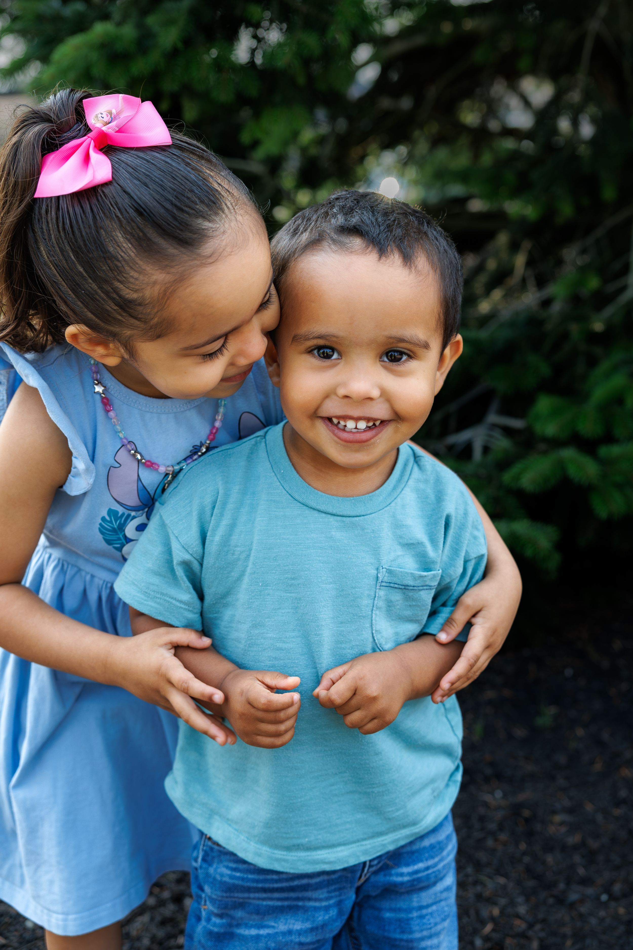 A young girl with a pink bow kisses her smiling little brother outdoors near green trees.