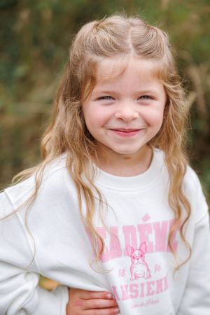 Happy young girl with long blonde hair wearing a white Academie Parisienne sweatshirt outdoors.