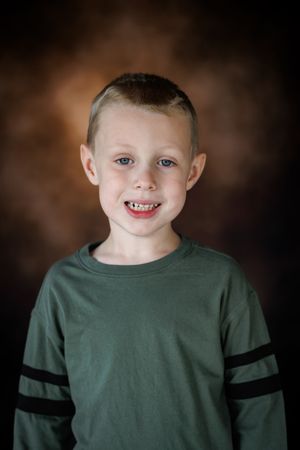 Smiling young boy with blue eyes wearing a green long-sleeve shirt posed against a brown studio backdrop.