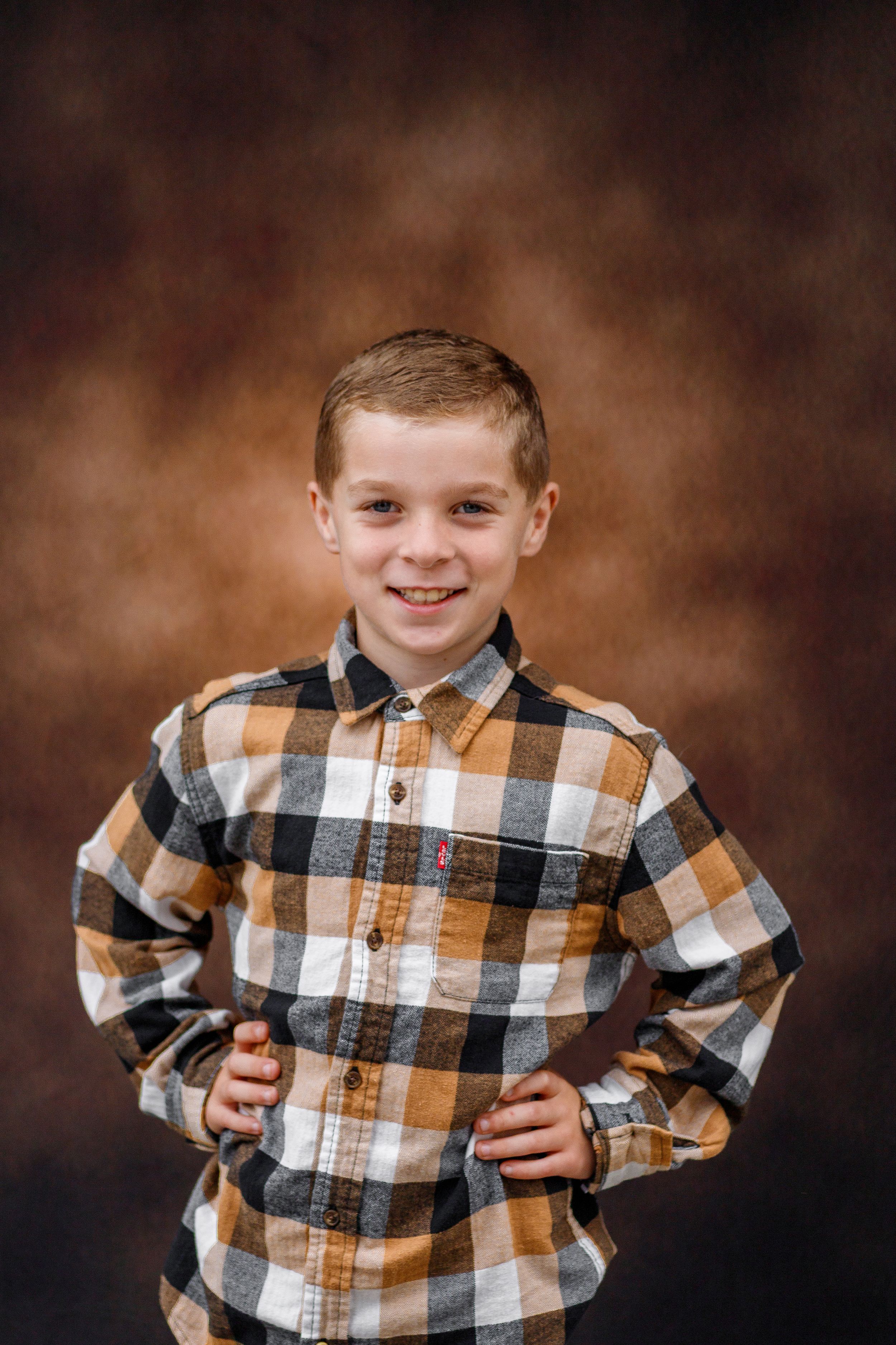 Smiling boy in plaid Levi's shirt posing with hands on hips against a brown studio backdrop.
