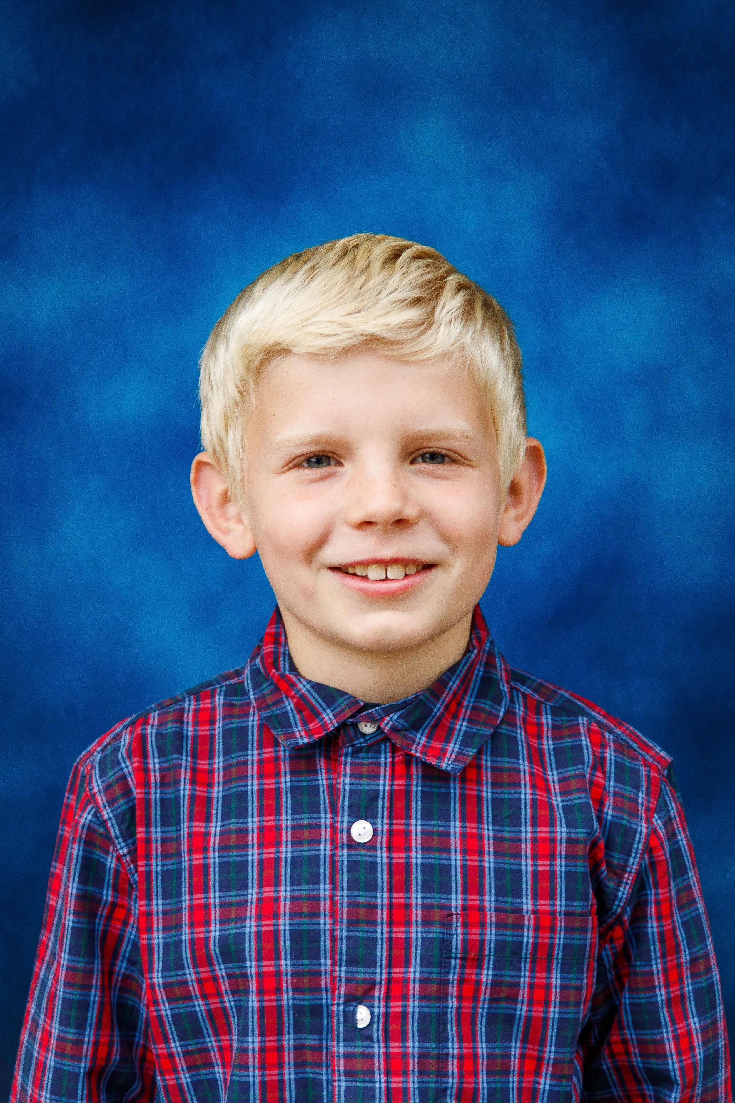 Smiling blonde boy in a red plaid shirt poses for a school portrait against a blue background.