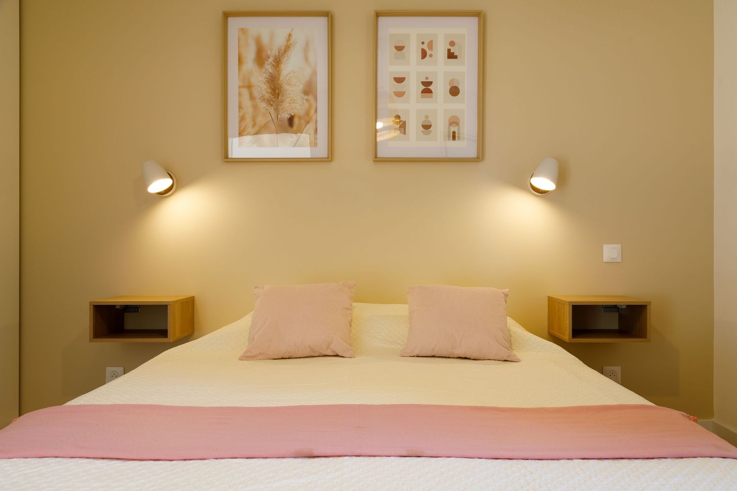 Chambre d'hotel chaleureuse avec un lit double, des oreillers roses, des appliques murales à la lumière douce et deux gravures botaniques.