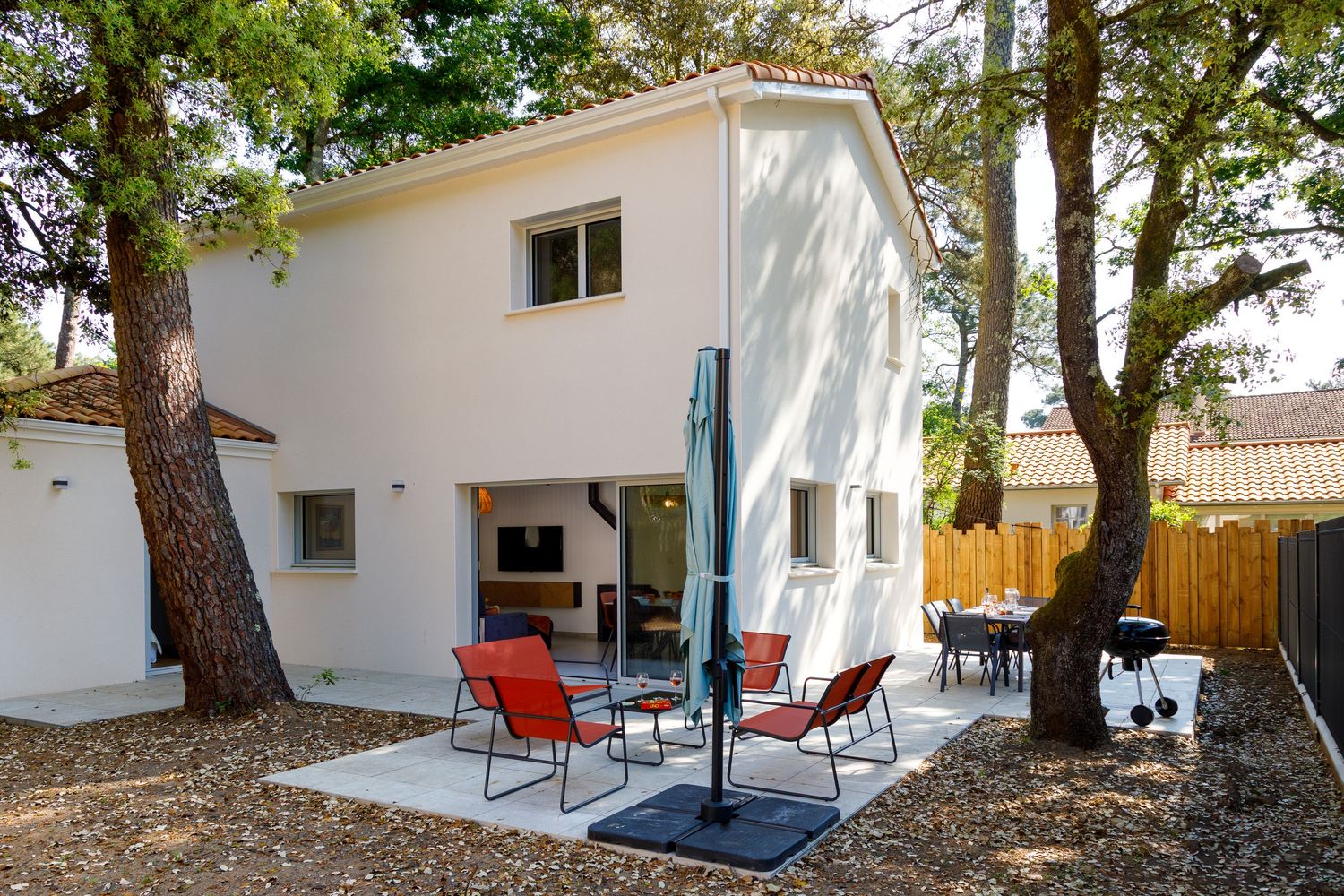 Maison blanche à deux étages avec terrasse dotée de chaises rouges et d'un parasol, ainsi que d'arbres matures dans un jardin paysager.